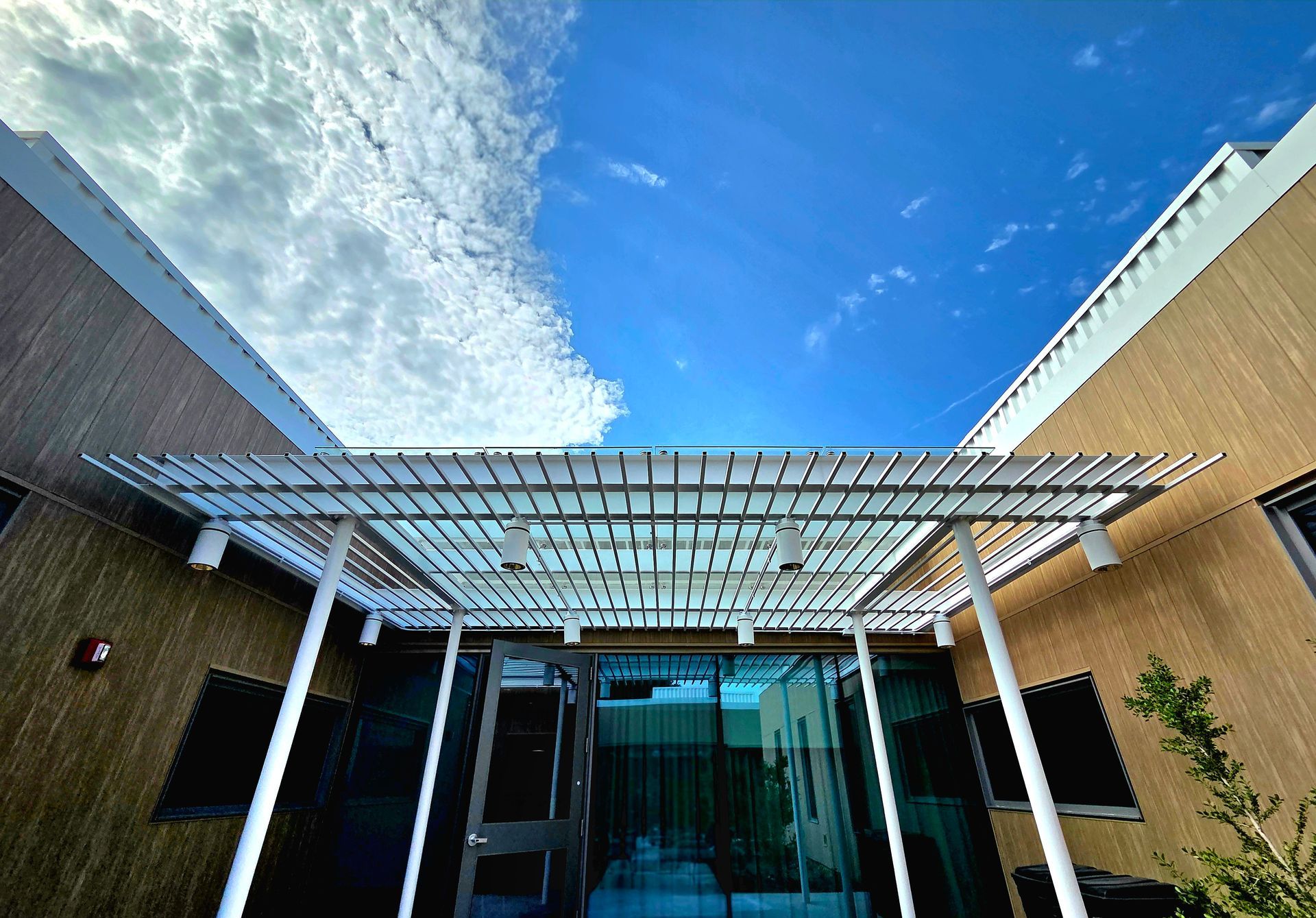 White pergola over a glass entrance, flanked by brown walls, against a blue sky with clouds.