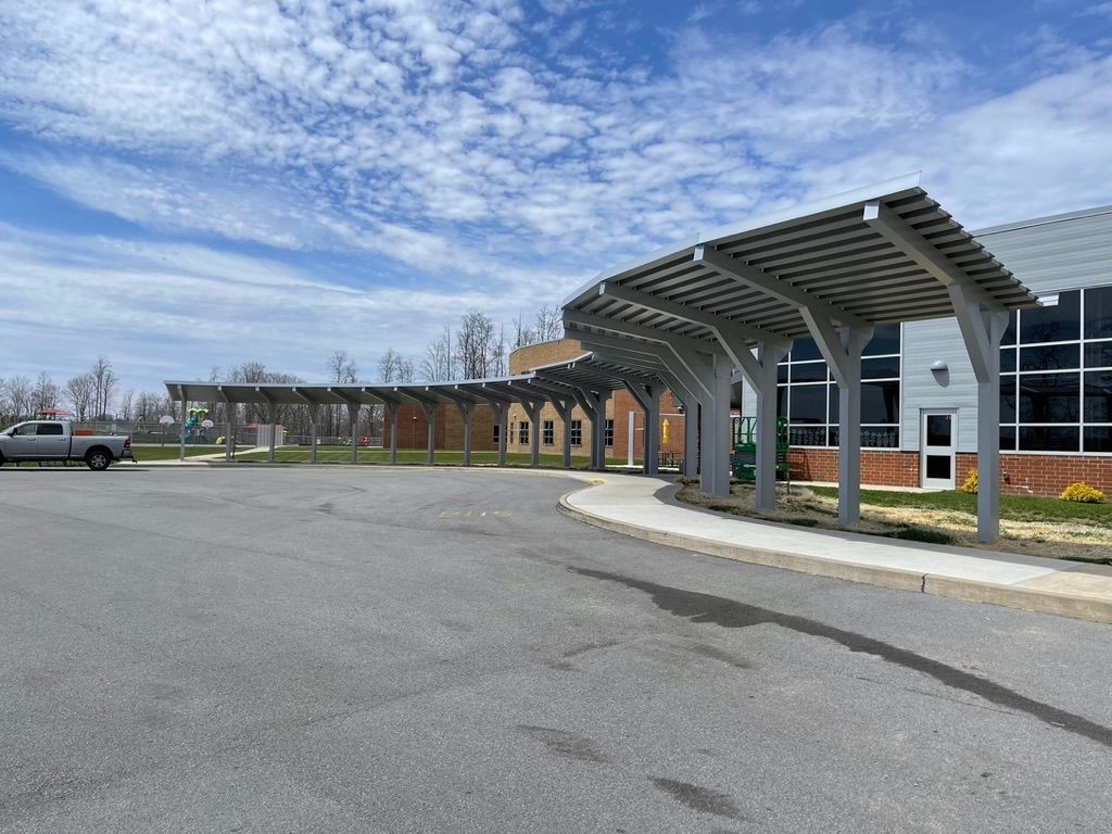 Canopy-covered walkway at a building with large windows and a gray facade, under a partly cloudy sky.