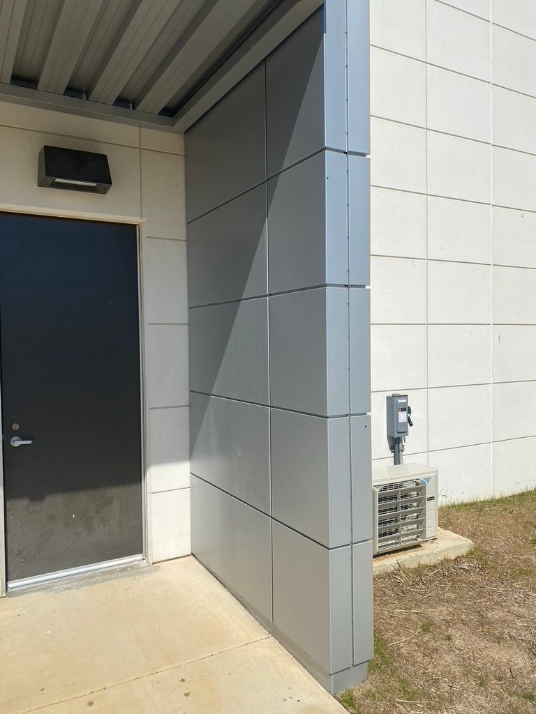 Corner of a building with a black door, silver panels, and an air conditioning unit.
