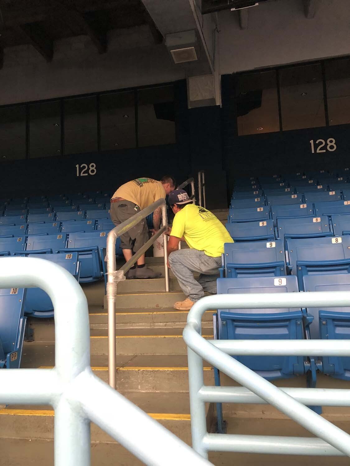 Two workers installing something on the steps inside an empty stadium. One is on the stairs, other is above.