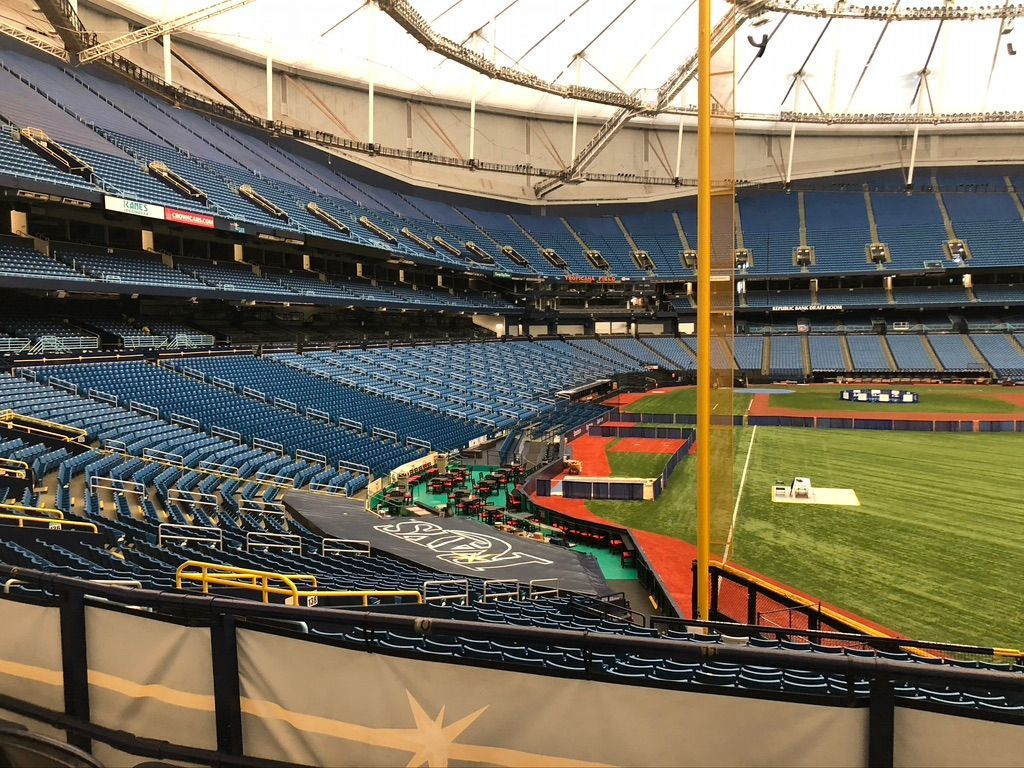 Interior view of a mostly empty baseball stadium with blue seats. The field is green, and the roof is white.