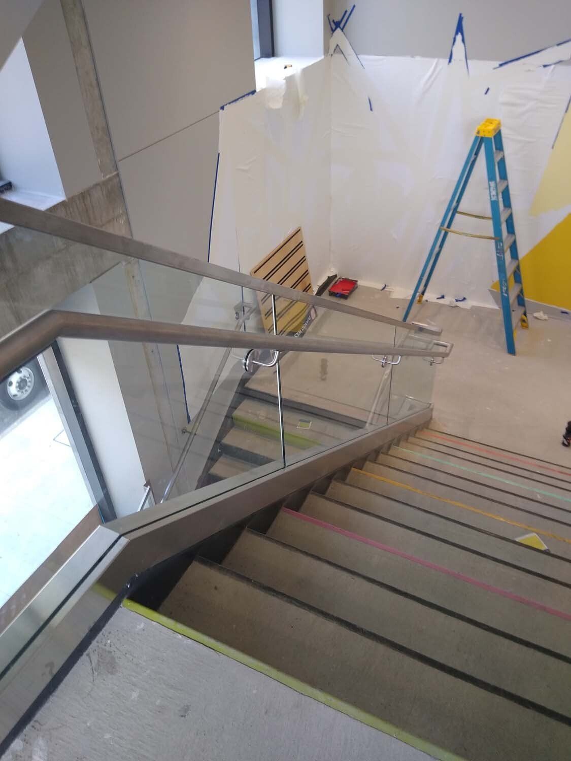 Staircase with glass railing, gray steps, and a blue ladder in a construction zone.