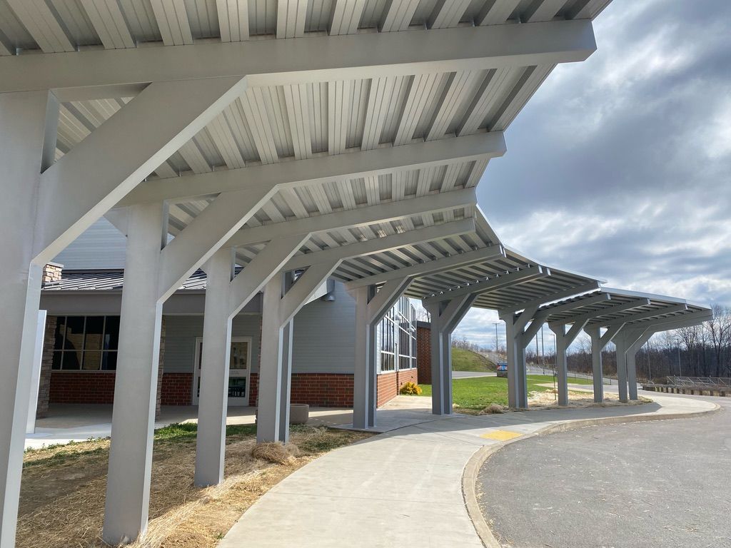 Covered walkway with grey supports, and curved roof. Paved path leads along a building with a cloudy sky overhead.