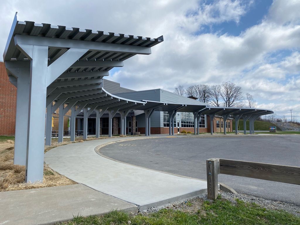 Gray-painted, curved pergola shelters a building entrance; a paved walkway leads to the door. Sunny day.