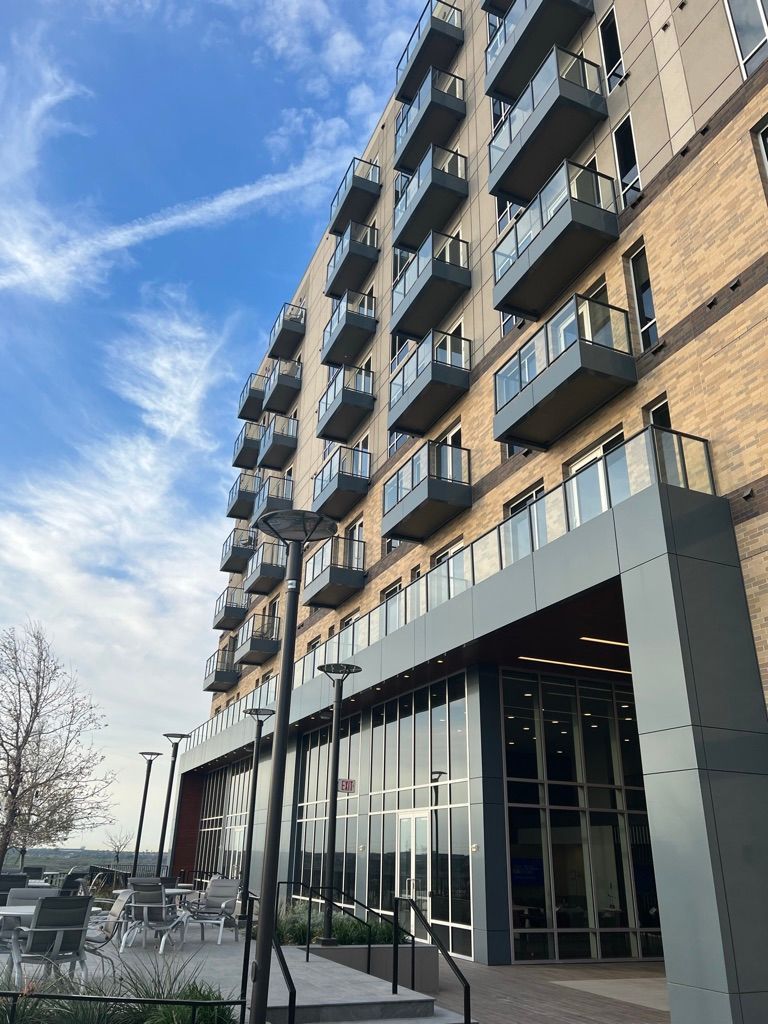 Modern apartment building with balconies, glass entrance, and outdoor seating under a cloudy blue sky.