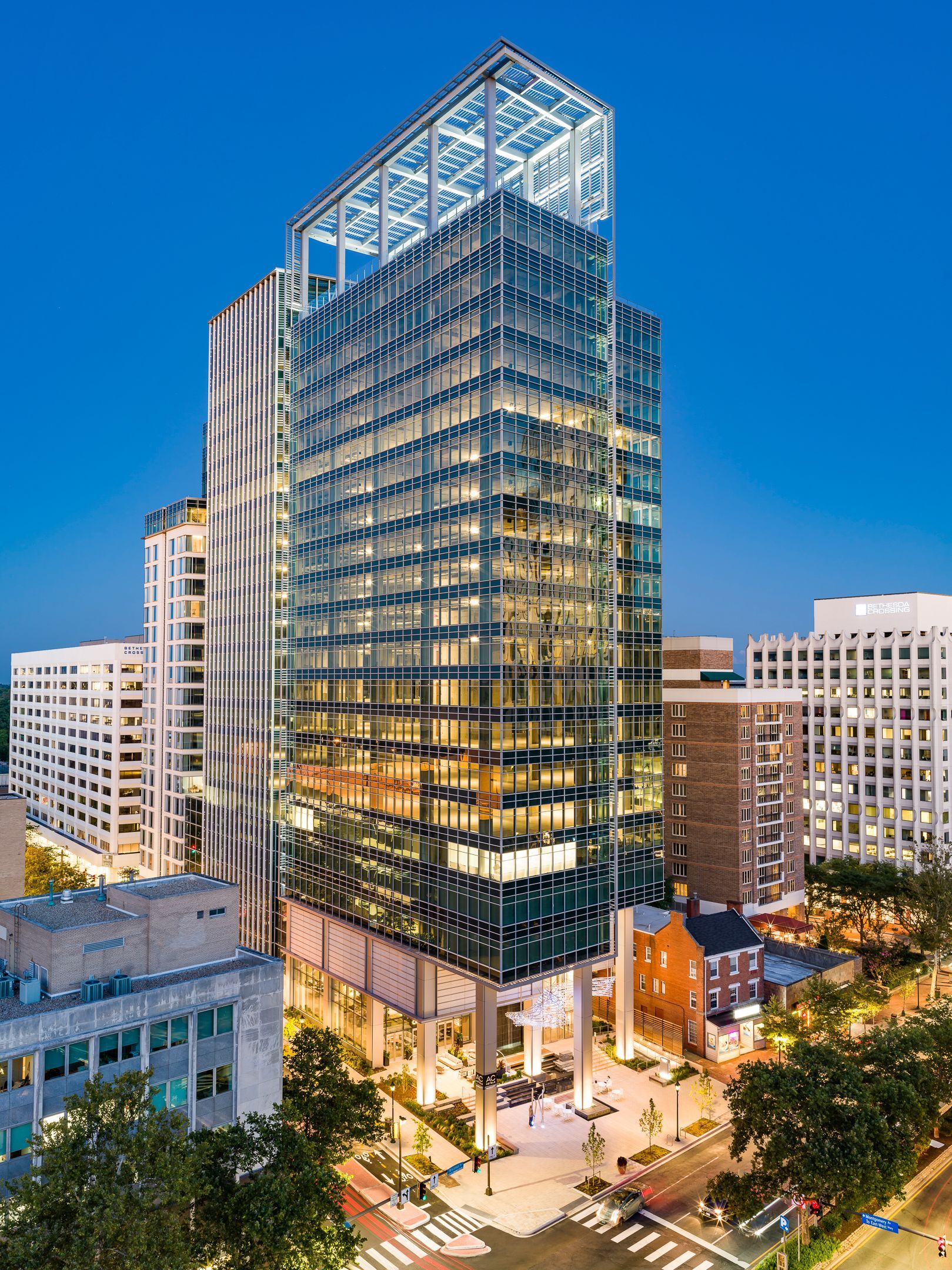 Modern glass skyscraper with open top, lit at dusk, in an urban environment with other buildings and a street below.