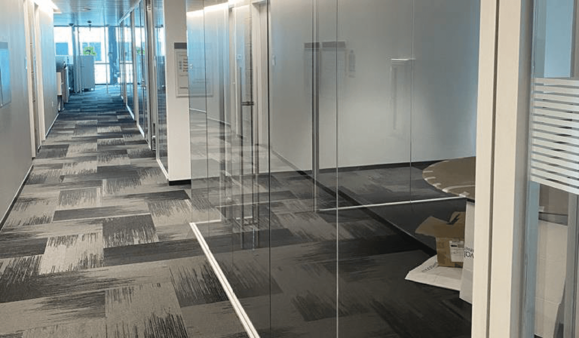Office hallway with glass-walled rooms and patterned carpet. Fluorescent lighting and neutral tones.