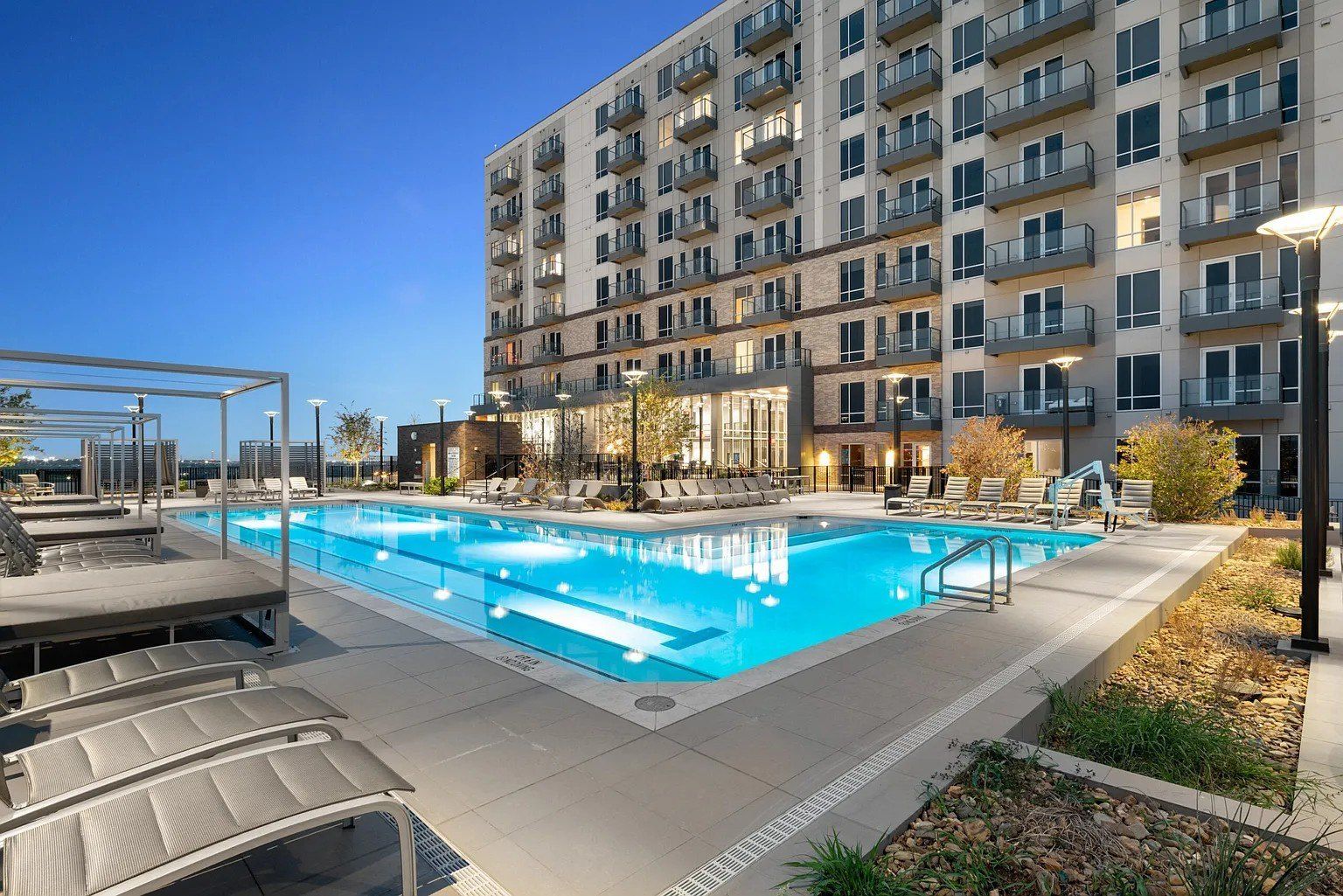 Swimming pool at dusk outside a multi-story apartment building. Lounge chairs and landscaping are visible.