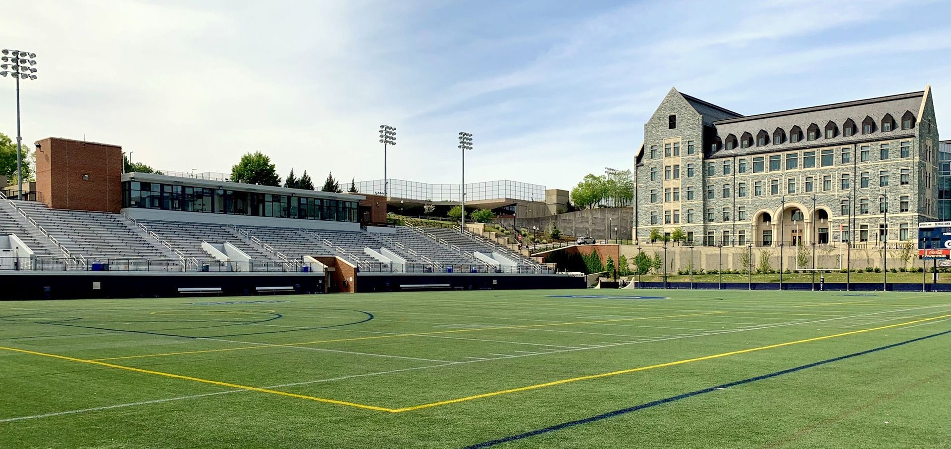 Football field with bleachers and a stone building on a sunny day.