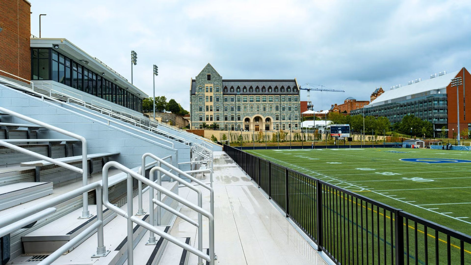 Bleachers and football field with large stone building in the background. Cloudy sky.