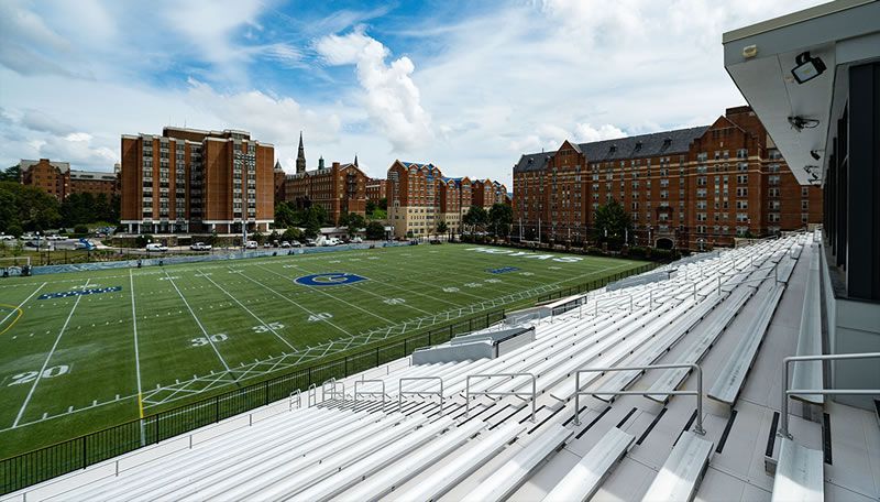 Football field with empty bleachers, surrounded by brick buildings under a cloudy sky.