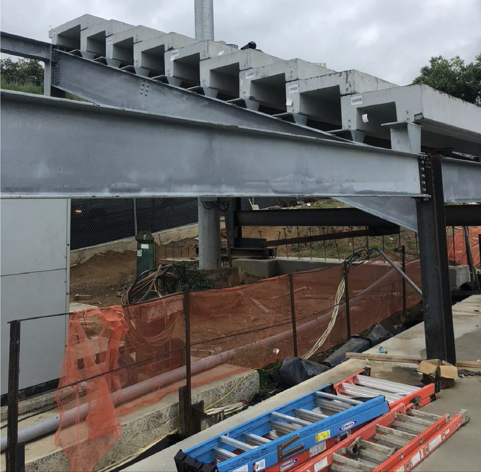 Construction site with gray steel beams and white concrete structures; orange safety fence.