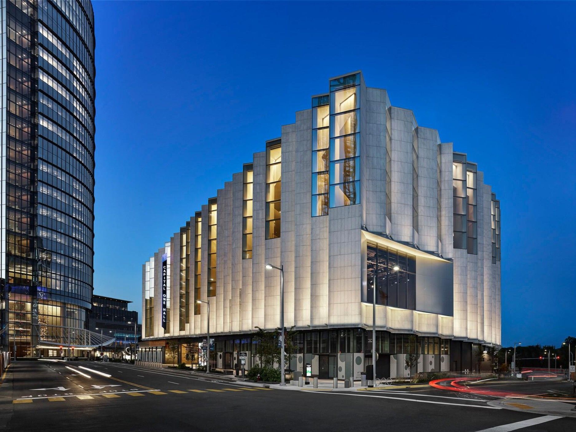 Modern building with illuminated exterior, at dusk, next to a tall glass tower. Street and traffic lights.