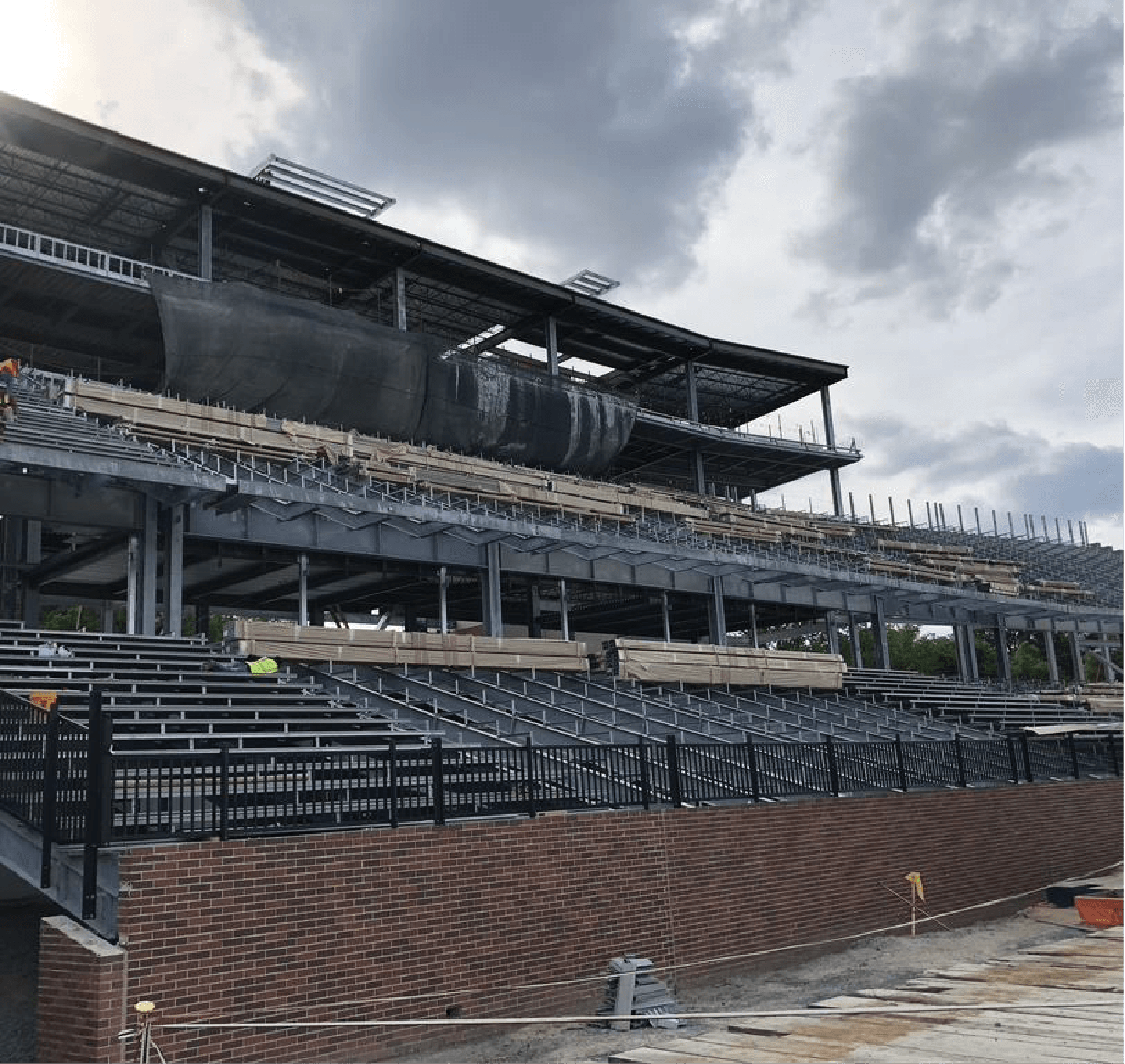 Construction of a multi-tiered stadium with steel framing and brick base under an overcast sky.