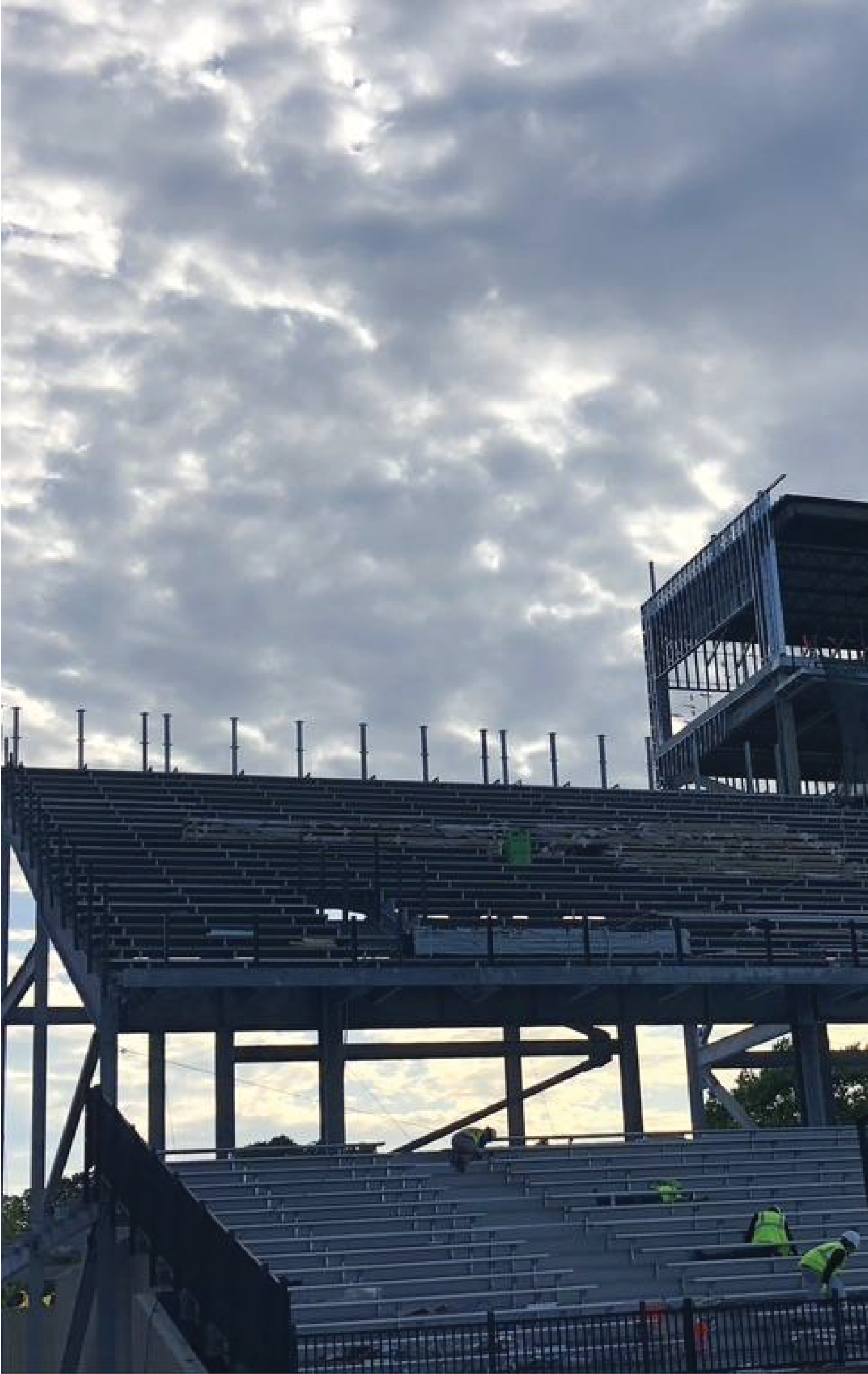 Construction site of a stadium under a cloudy sky. Workers in green vests are present.