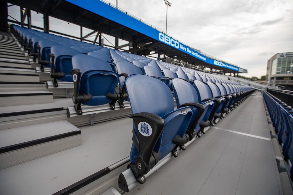 Empty blue stadium seats at an outdoor venue, under an overcast sky.