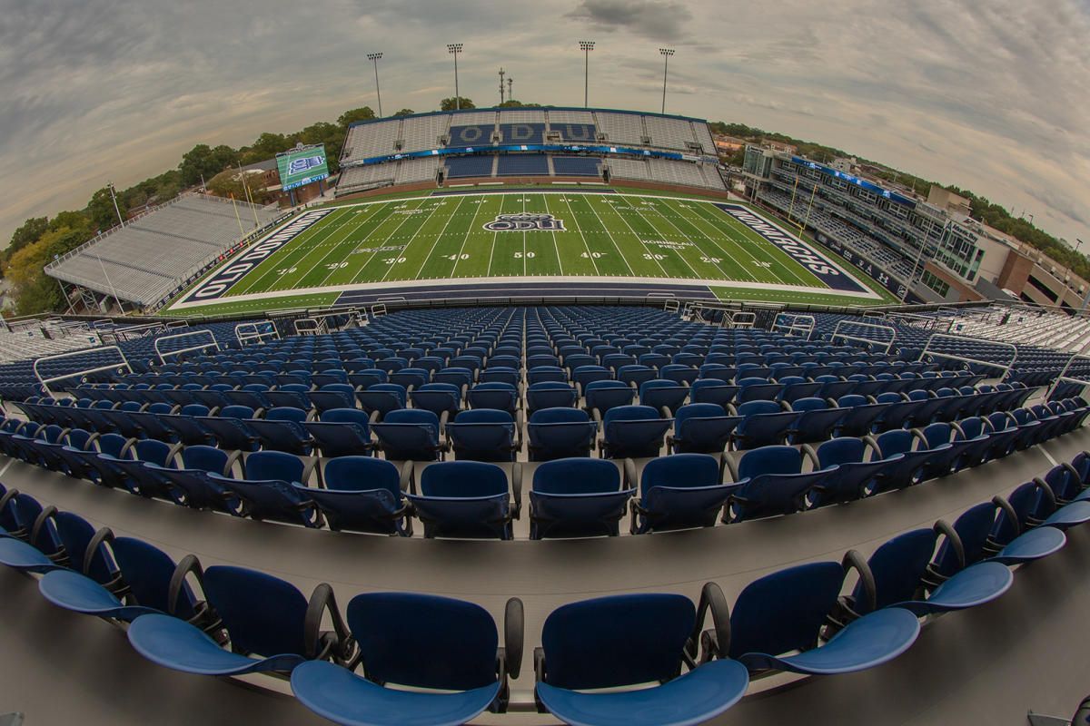Empty blue stadium seats overlooking a football field. The sky is overcast.