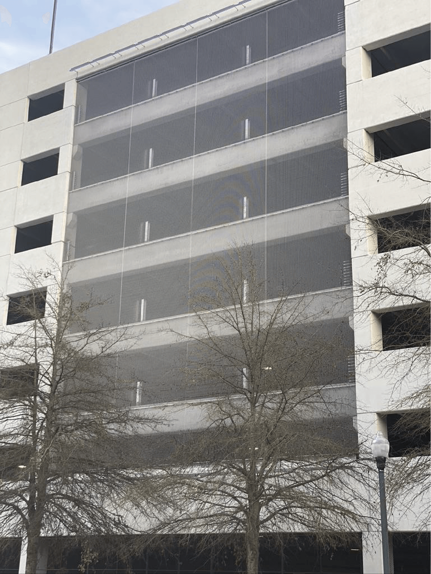 Multi-story parking garage exterior with metal mesh facade and bare trees in front.