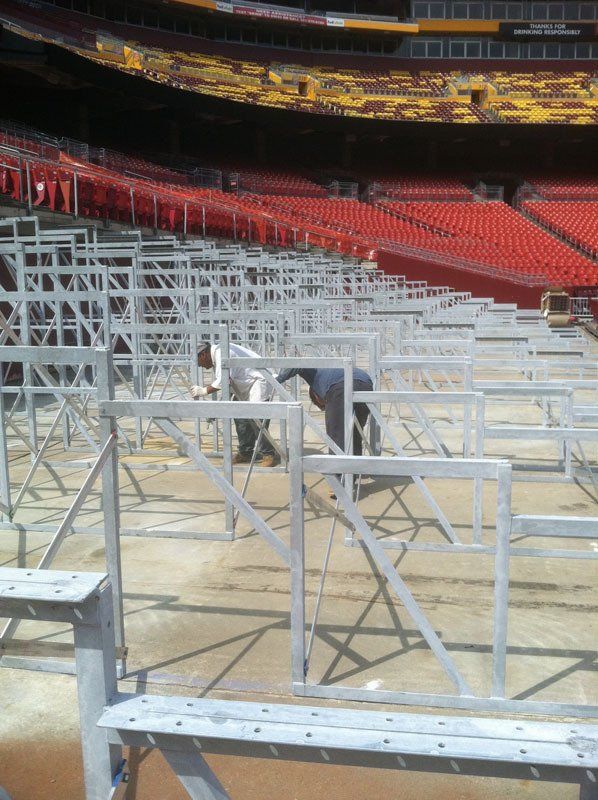 Workers assembling metal structures on a field in a stadium with red seating.