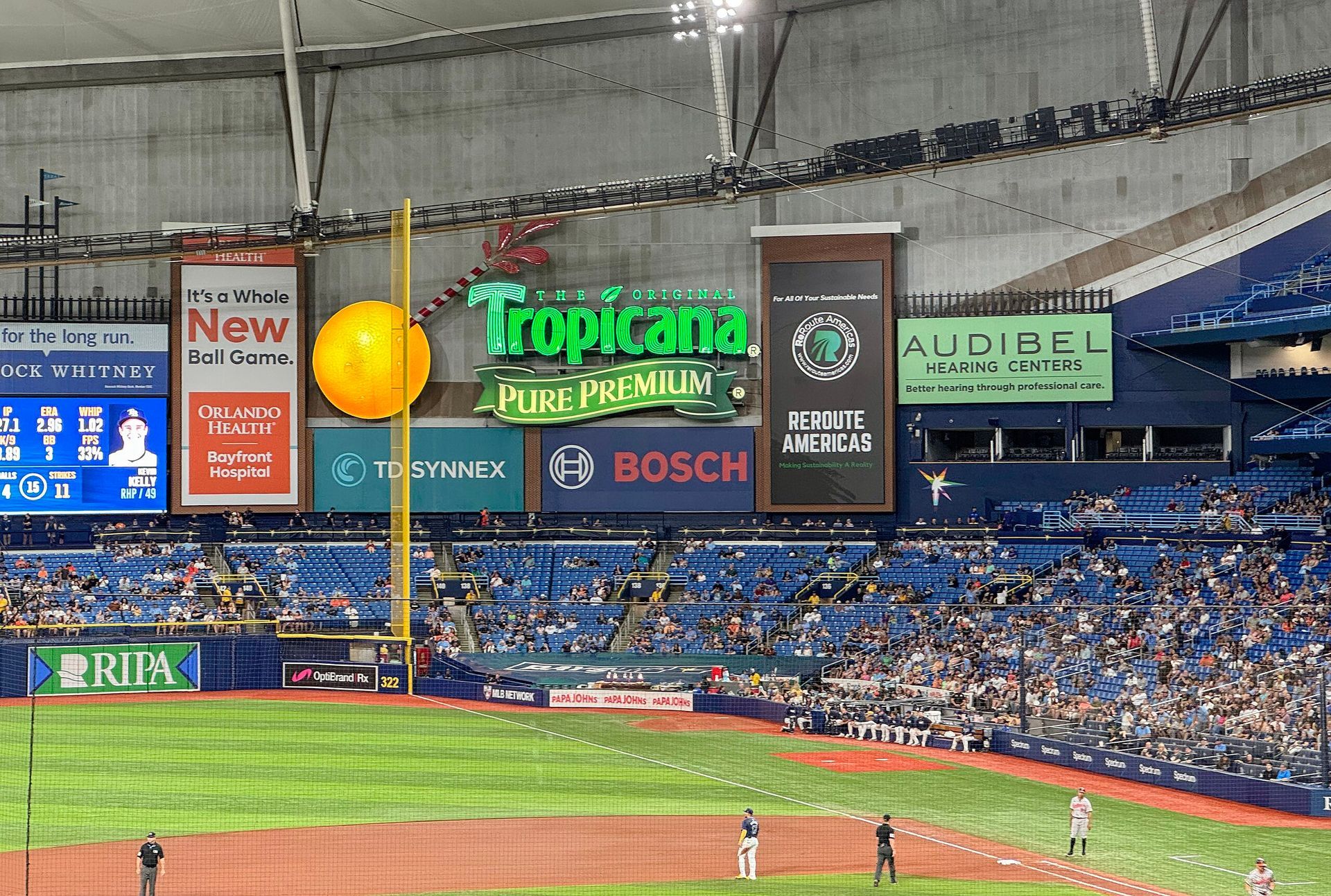 Baseball game at Tropicana Field, Tampa. Large scoreboard with advertisements, players on field, crowd in stands.