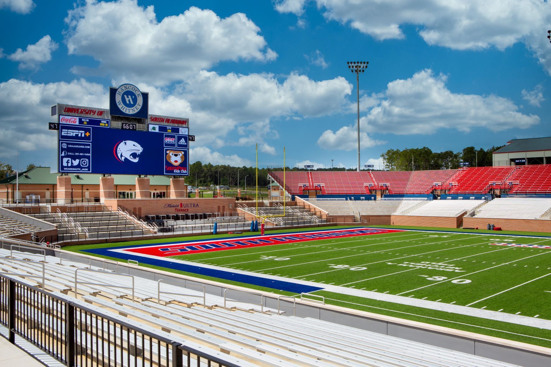 Football stadium with a large scoreboard, green field, and blue and red seating under a cloudy sky.