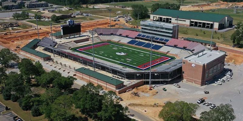 Aerial view of a stadium with a green field, red and blue seating, and a large scoreboard.