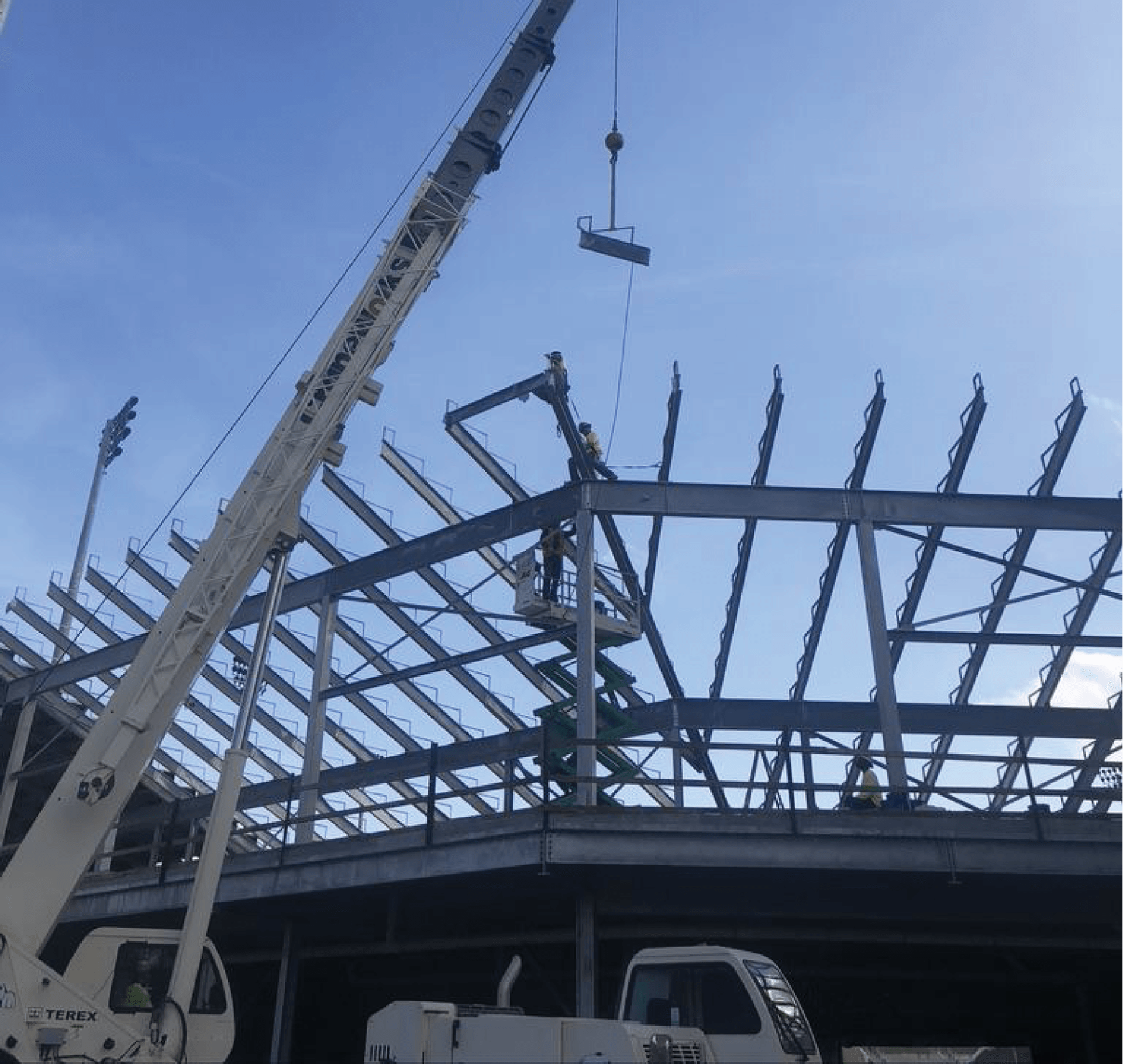 Crane lifting a steel beam for roof construction. Workers on a platform, clear blue sky.