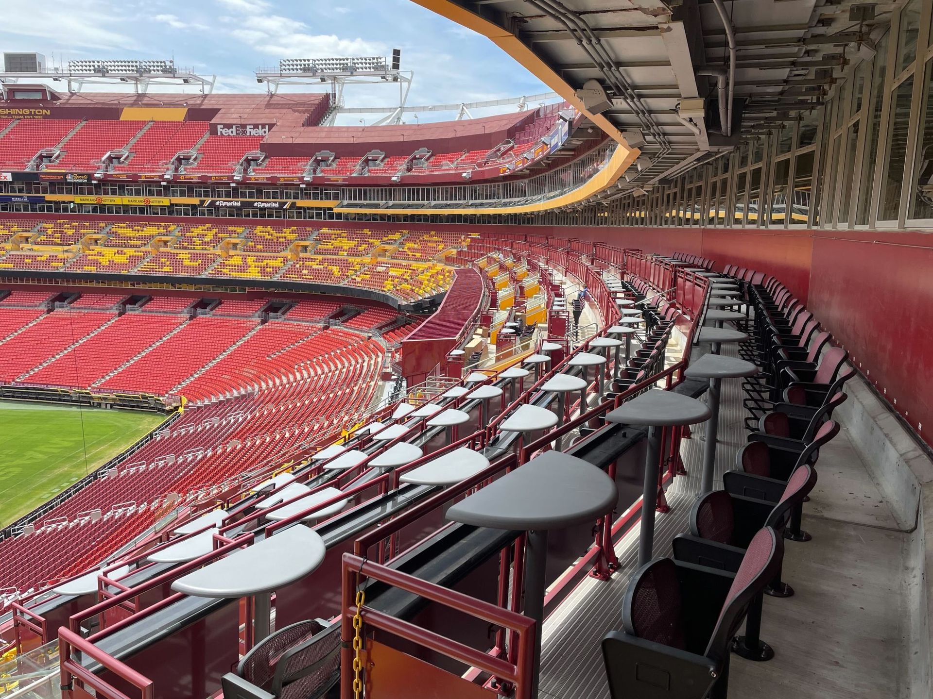 Empty stadium seating with red and yellow chairs, tables, and a green field.