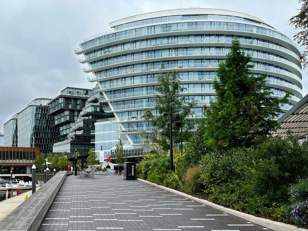 A modern waterfront building with curved design, walkway, trees, and cloudy sky in the background.