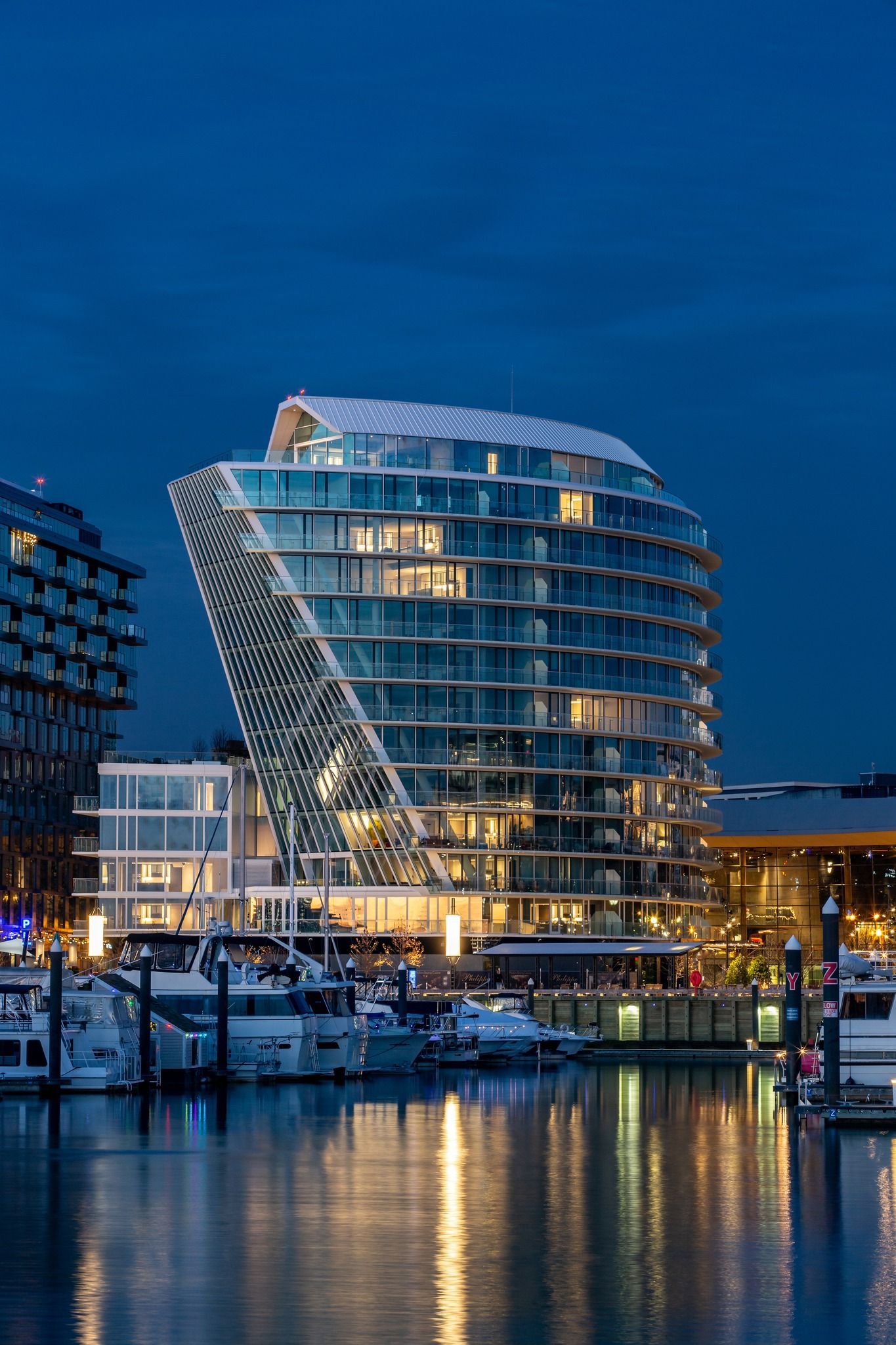 Modern, curved glass building with lit windows overlooks a marina with docked boats at dusk.
