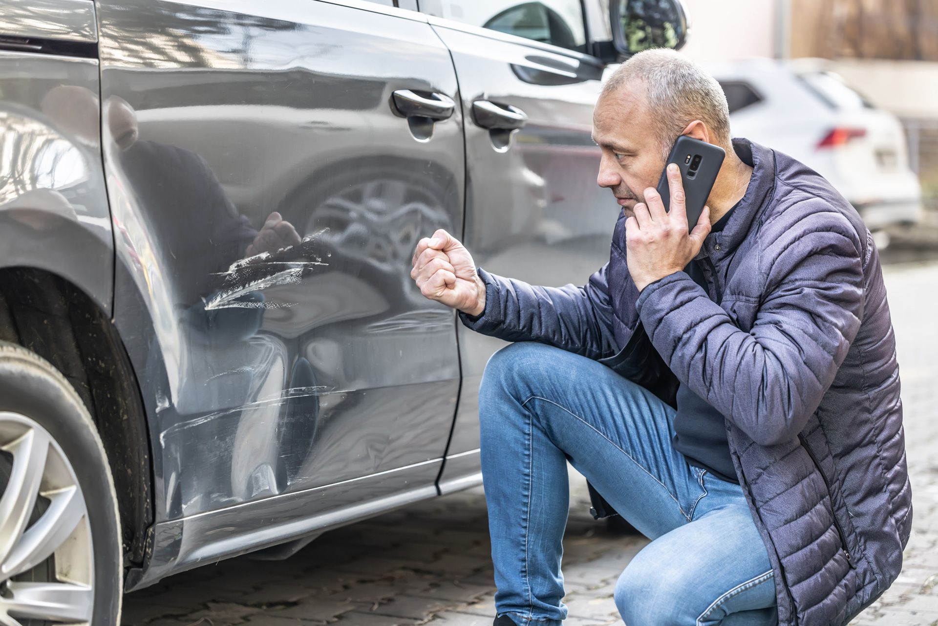 Man on phone, kneeling beside a dented gray car on a city street. He is upset, pointing at the damage.