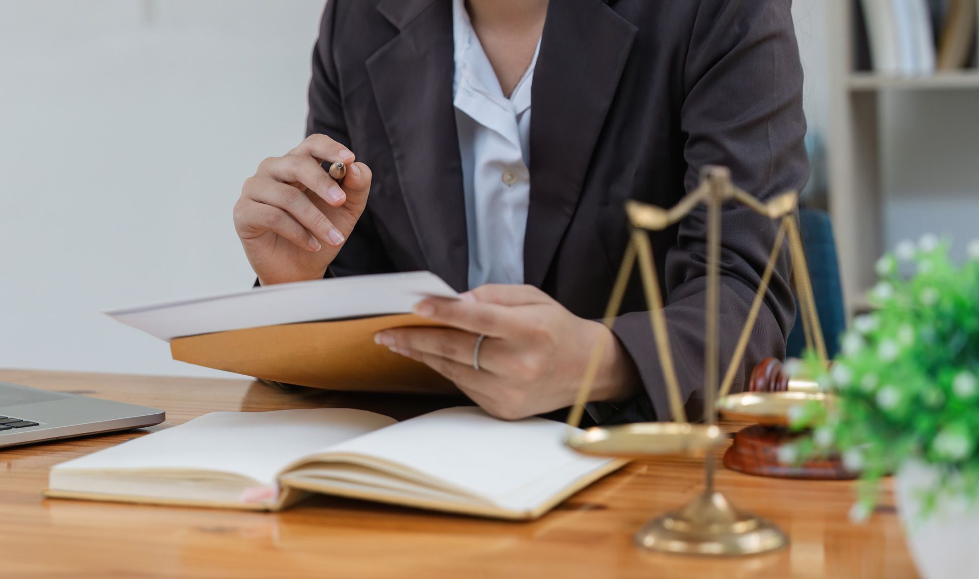 Person in a blazer examines documents at a desk with a scale of justice and open book.