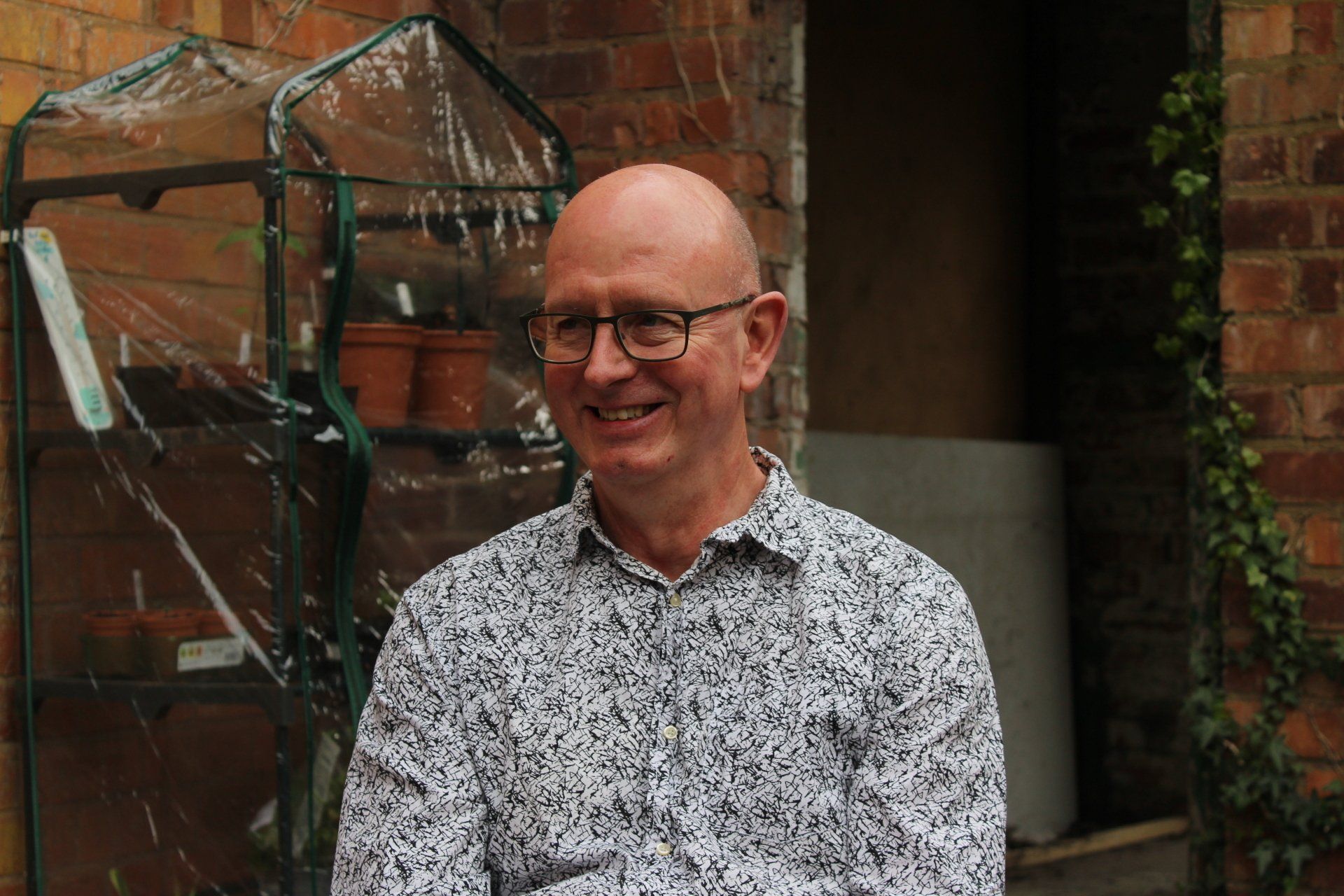 Man with glasses smiles, wearing a patterned shirt, in a garden.