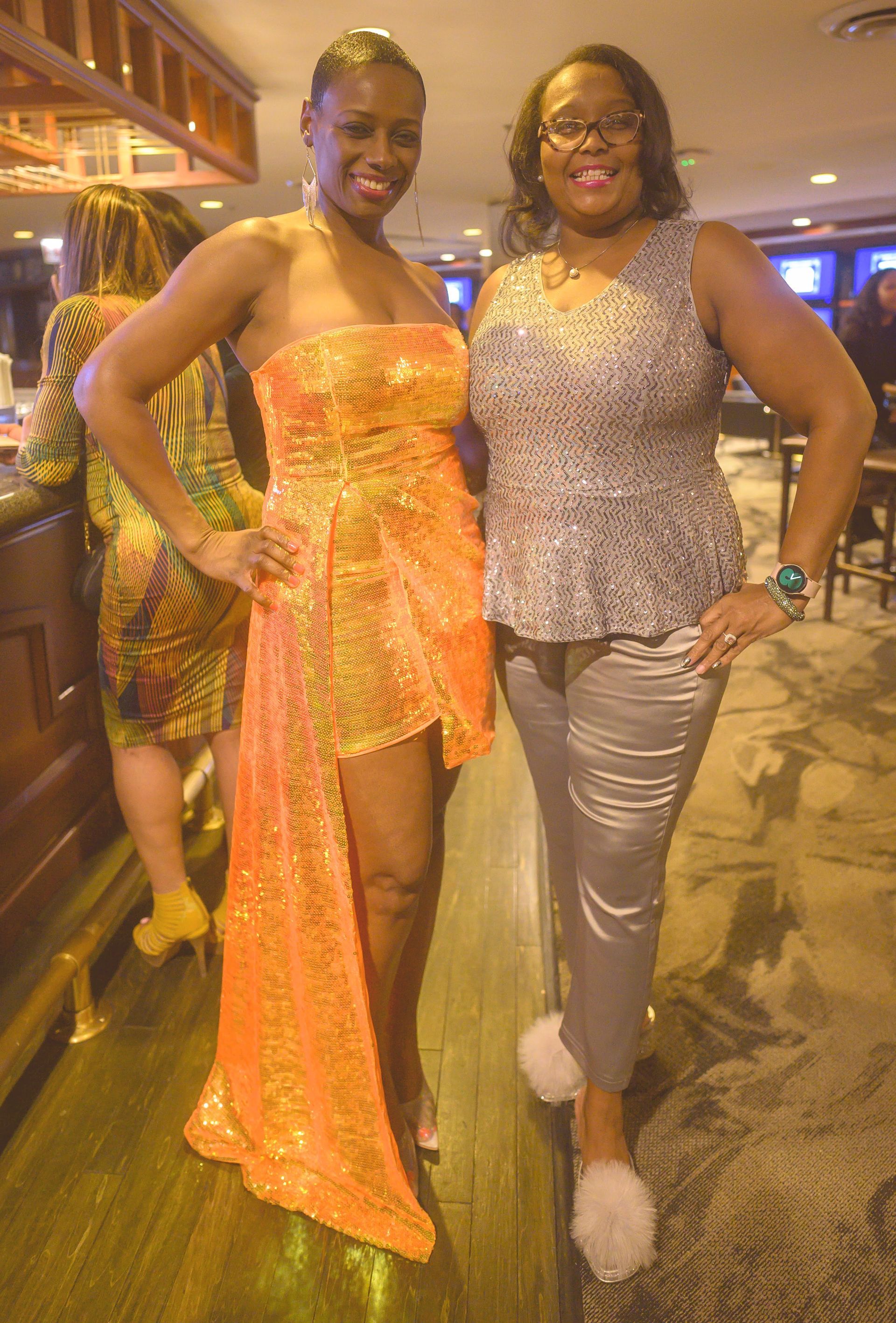 Two women are posing for a picture together in a room . one of the women is wearing an orange dress.