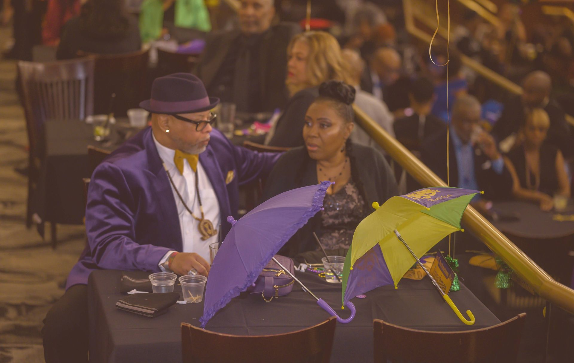 A man in a purple suit is holding a purple umbrella while sitting at a table with a woman.