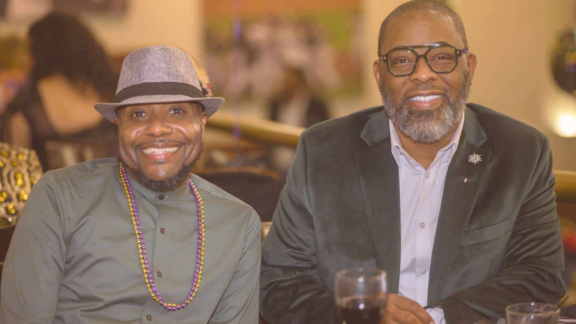 Two men are sitting next to each other at a table with glasses of wine.
