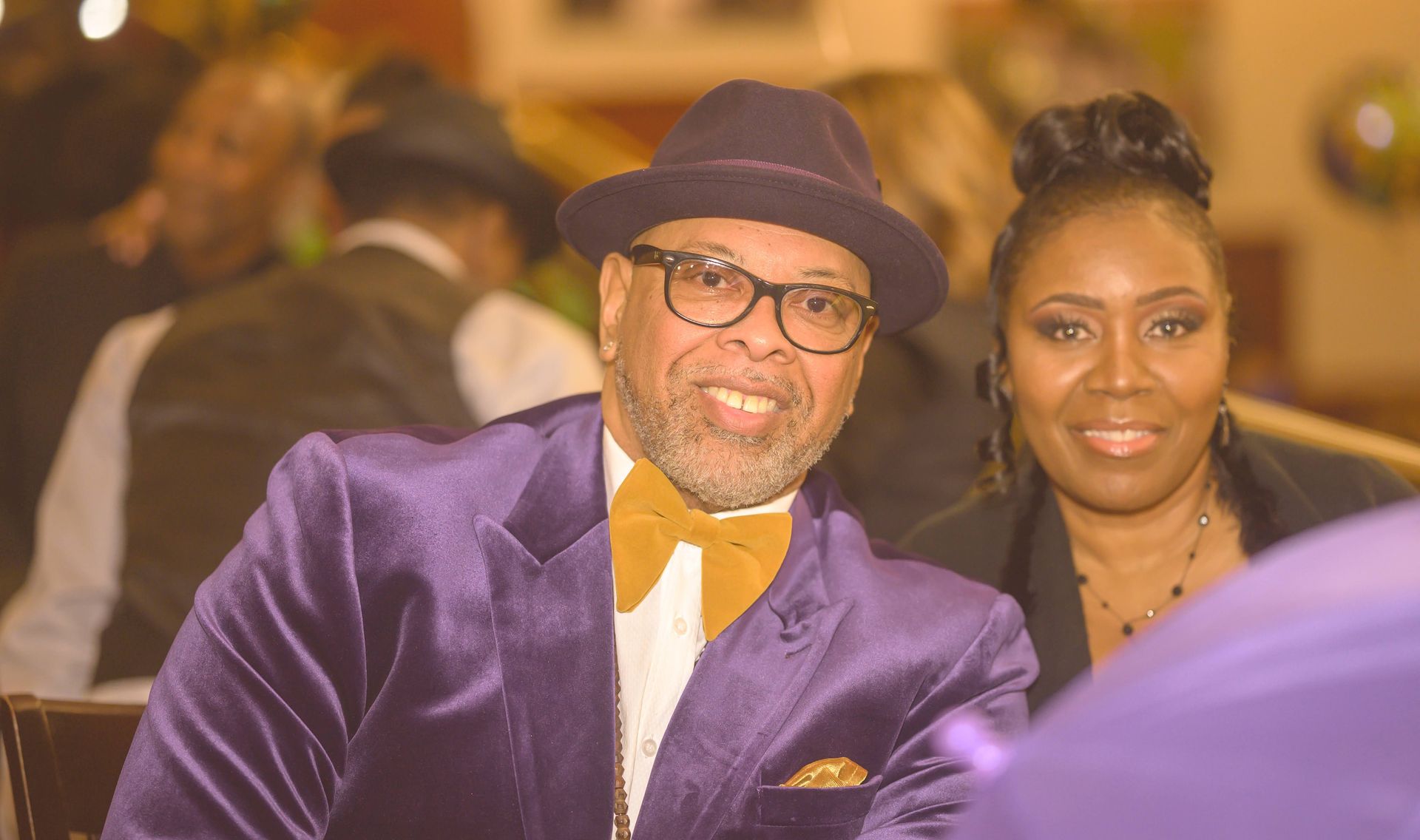 A man in a purple suit and hat is sitting next to a woman.