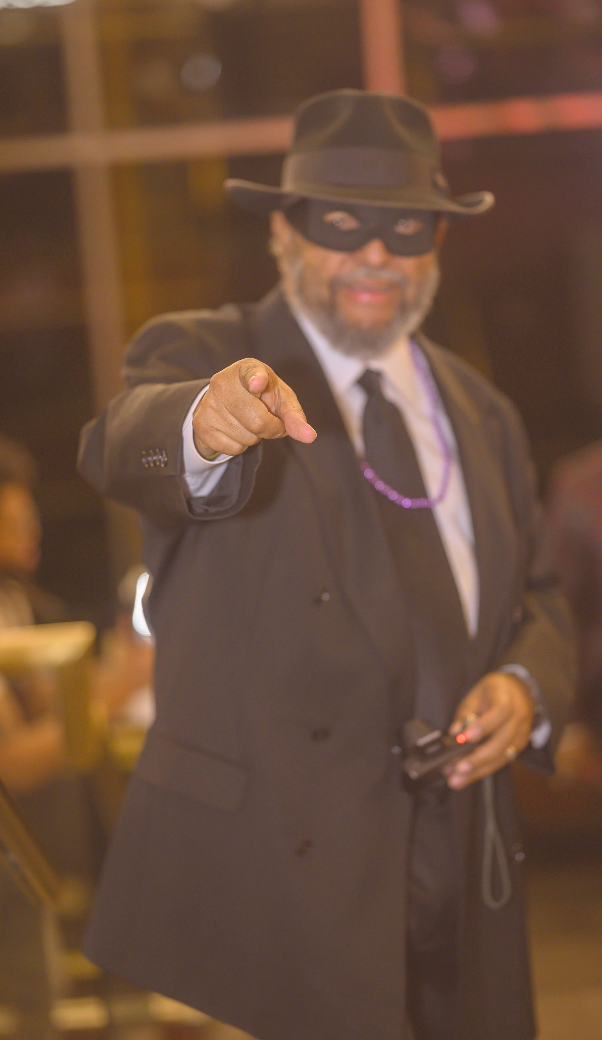 A man in a suit and mask is pointing at the camera.