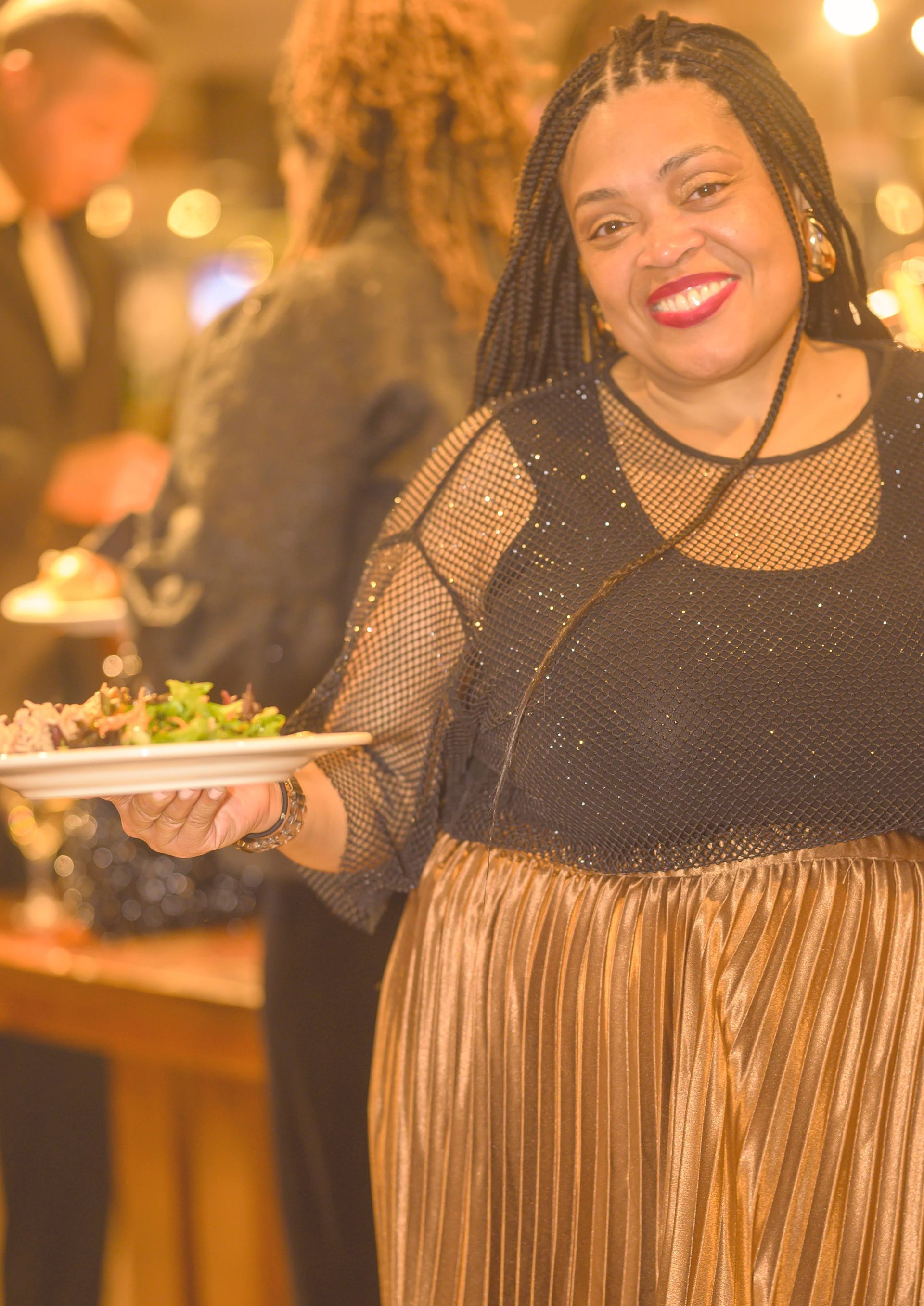 A woman in a black top and gold skirt is holding a plate of food.