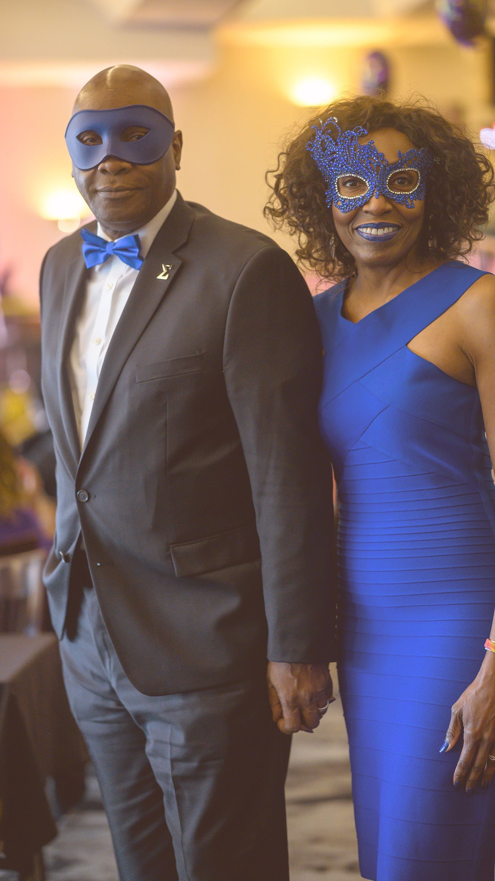 A man in a suit and a woman in a blue dress are holding hands and wearing masks.