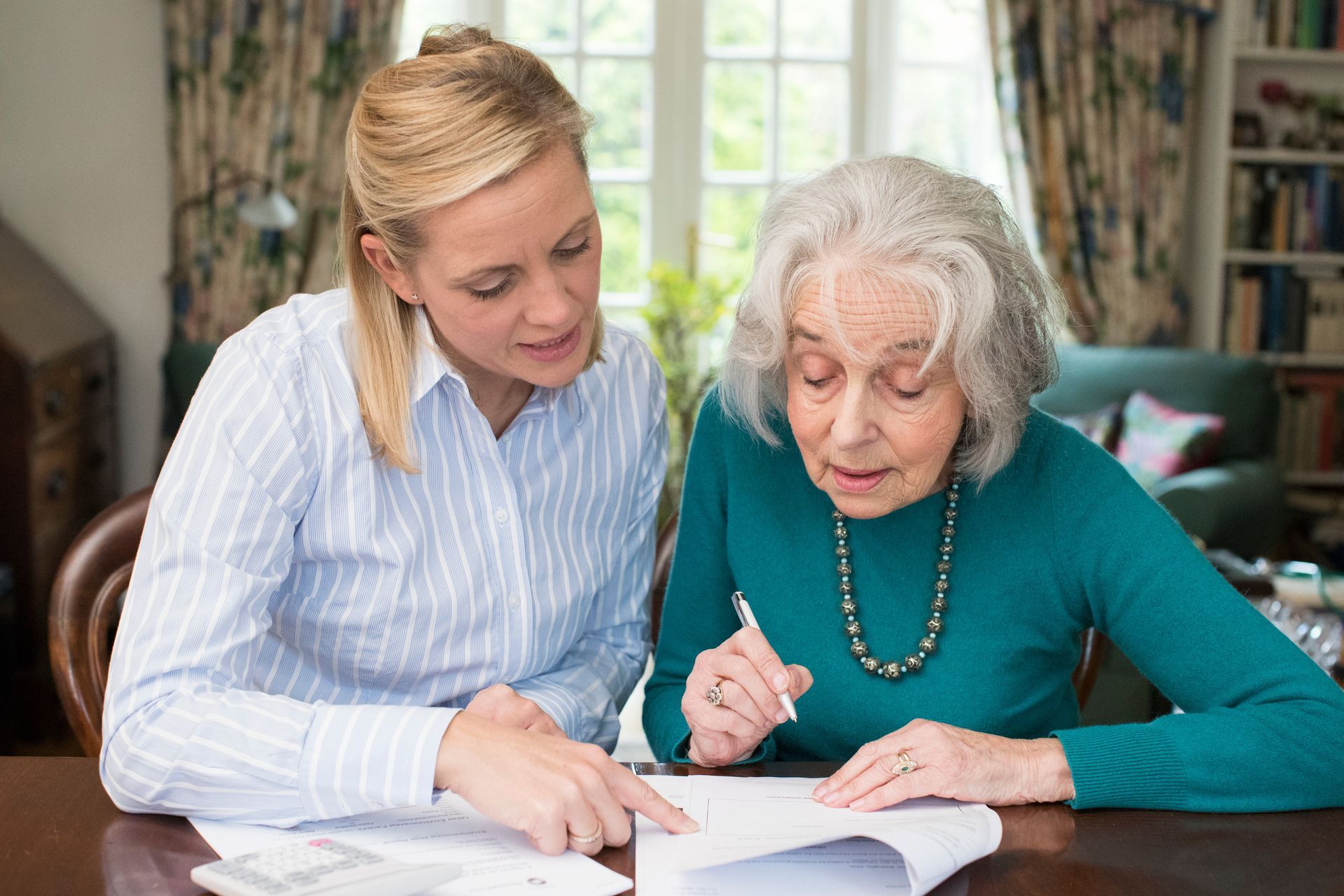 A person in a striped shirt points to a document while another person sits nearby holding a pen at a wooden table.