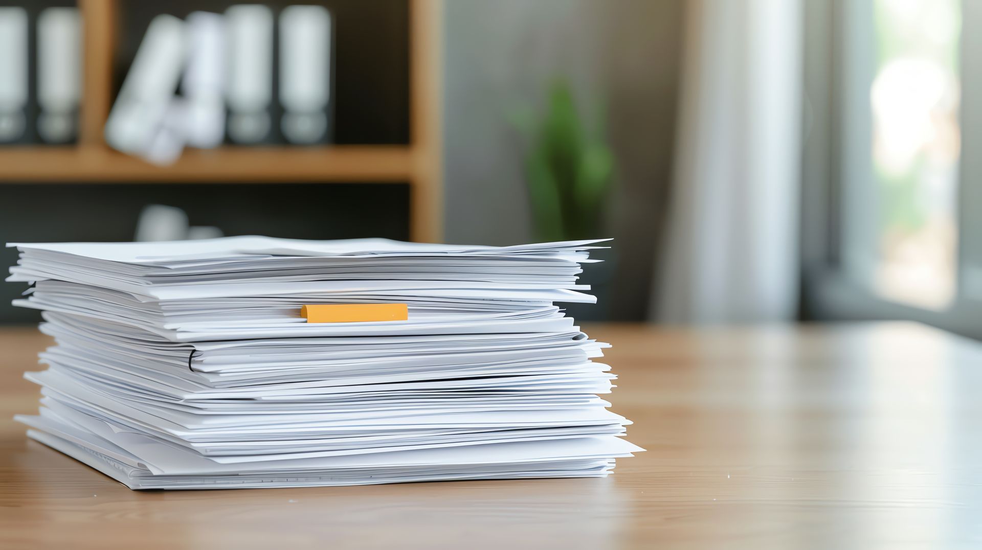 A tall stack of white paper with a small orange sticky note sits on a wooden desk in an office setting.
