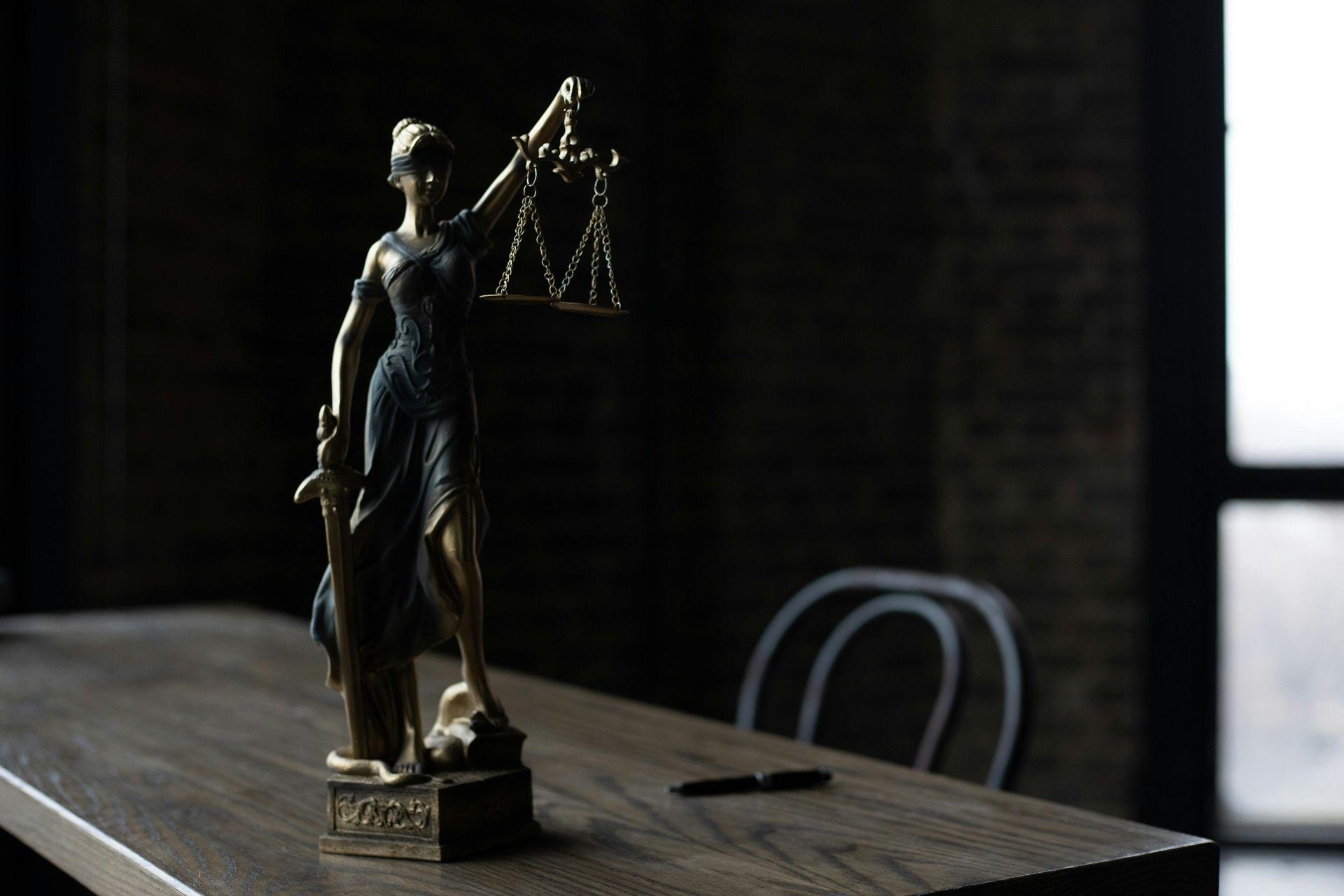 A bronze statue of Lady Justice holding scales and a sword stands on a wooden desk with a pen and chair in the background.