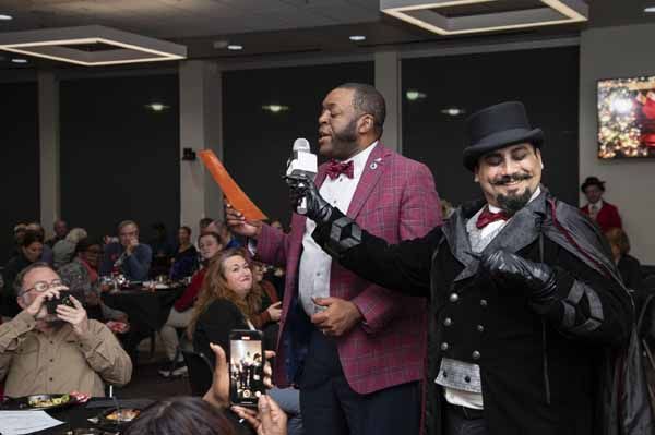 Man in blazer holds microphone, talks to magician in top hat. Event with guests.