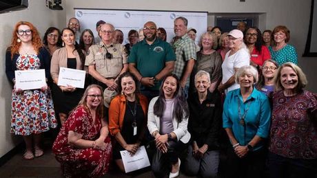 Group of smiling people posing indoors at a conference, some holding certificates, in front of a presentation screen.