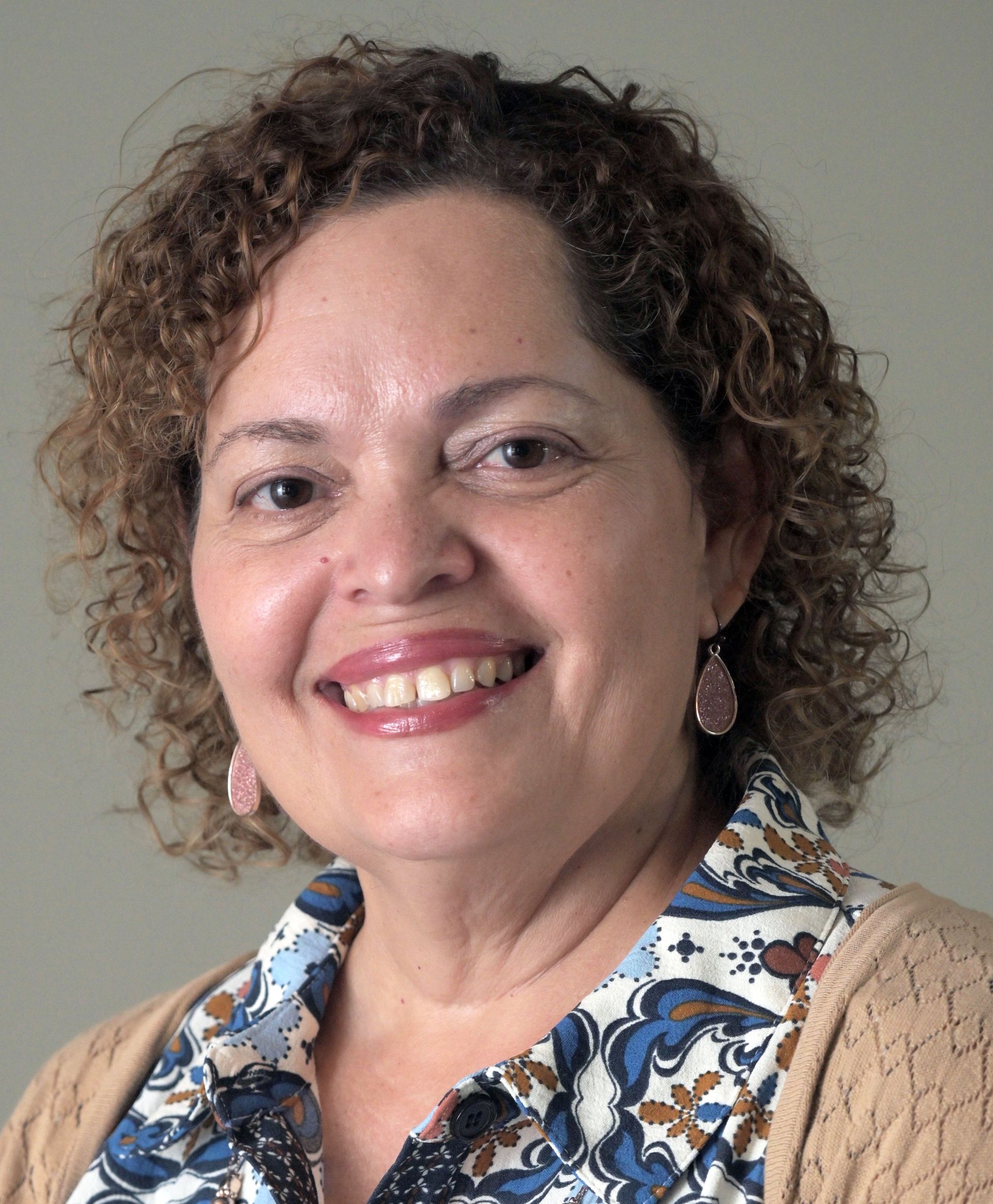 Woman with curly brown hair smiles, wearing a black and white patterned top, indoors.