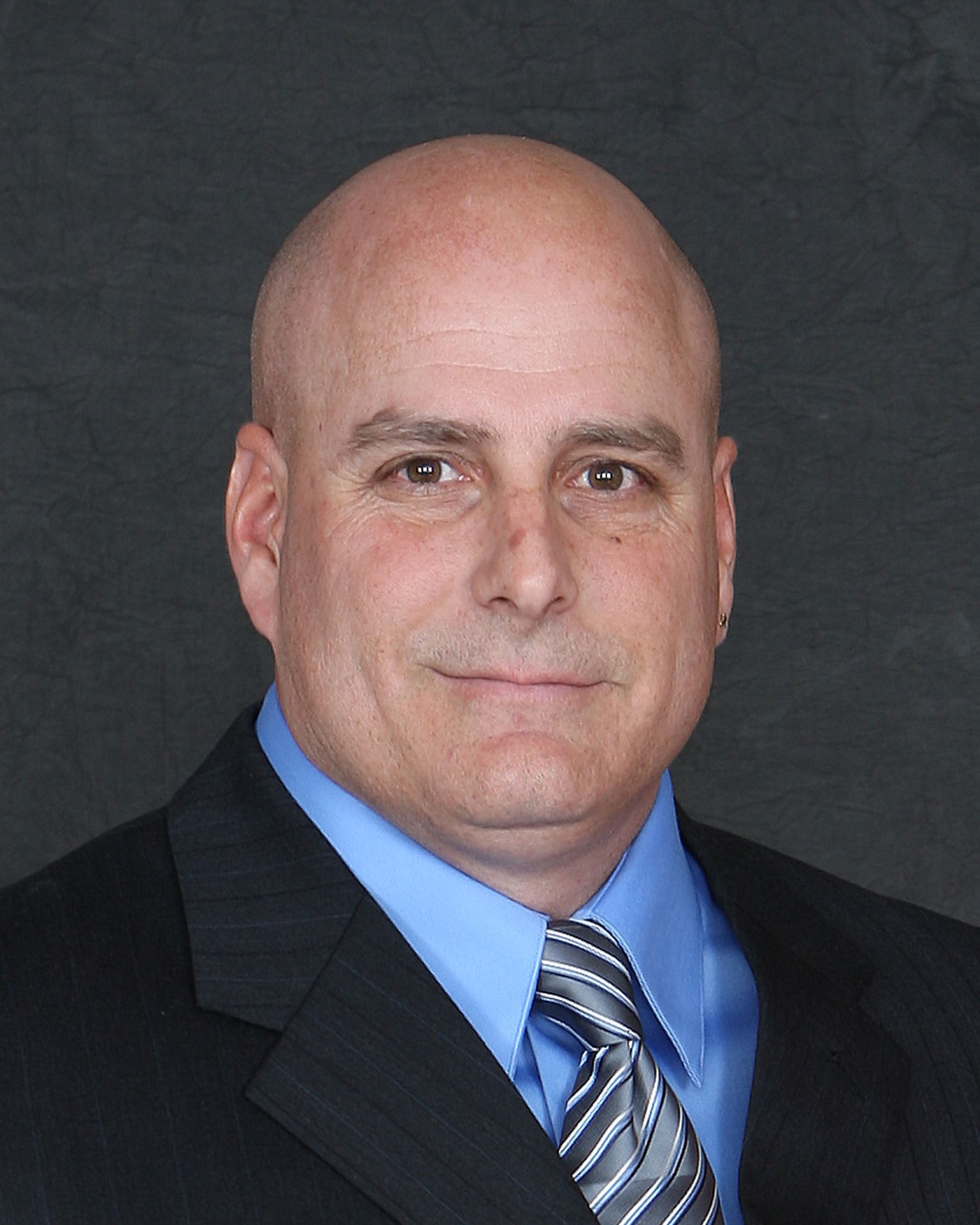 Bald man in a suit and tie, smiling slightly. Formal portrait against a gray background.