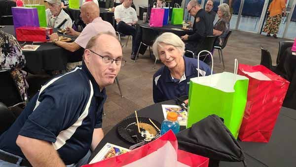 Two people at a table, smiling, surrounded by gift bags, indoors.