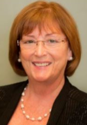 Woman with auburn hair, glasses, smiling, wearing pearls and a black jacket.
