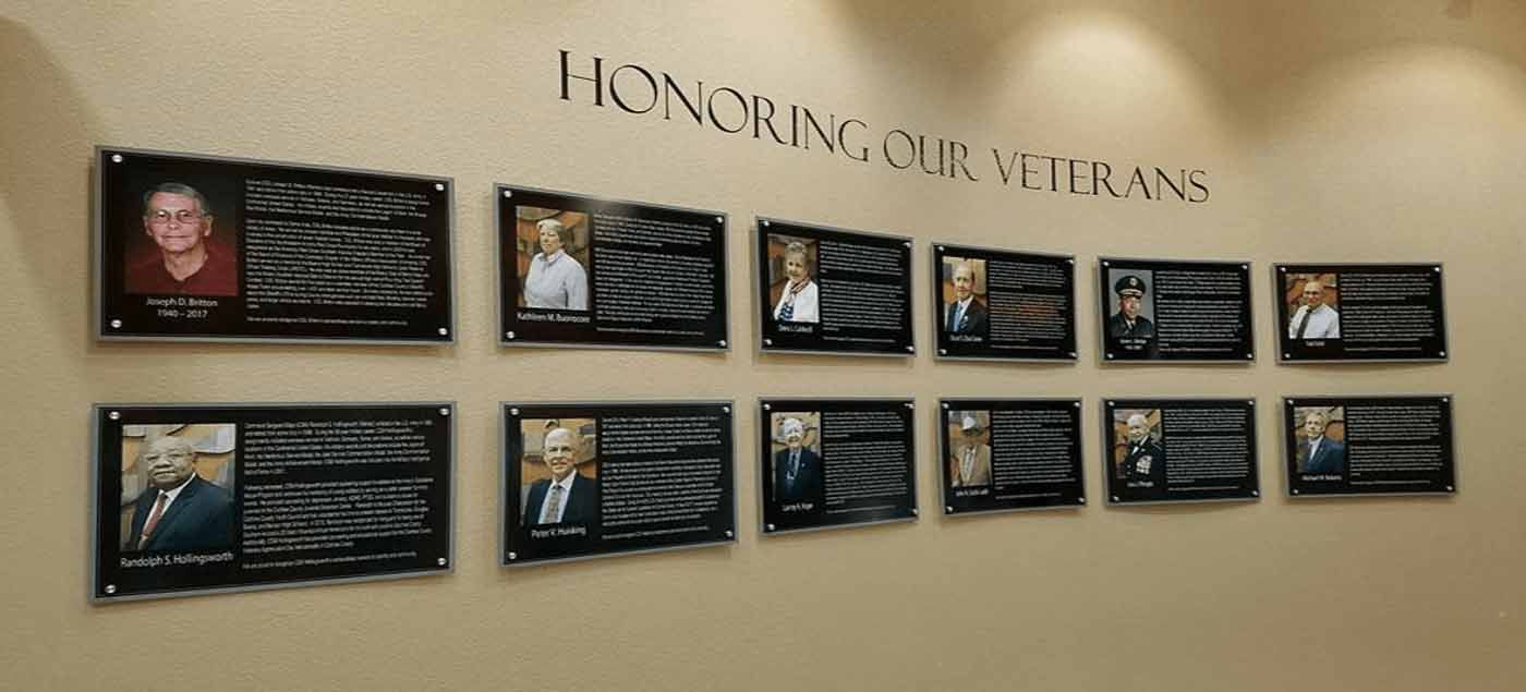 Wall with plaques honoring veterans. Black plaques with photos and text, mounted on beige wall. Text: 